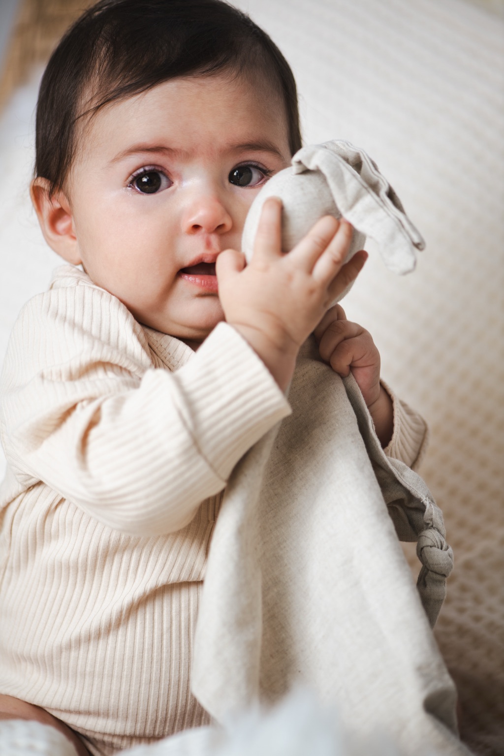 Dark eyed baby holding a knotted linen lovey to its mouth