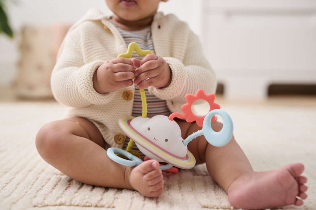 Baby in a cream cardigan on a rug playing with a smiling Saturn rattle