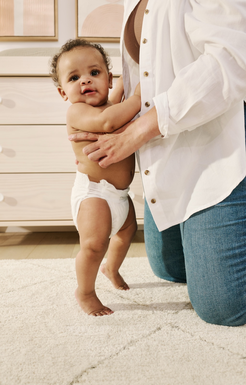 Diapered baby standing on a nursery rug holding a kneeling parent's arm