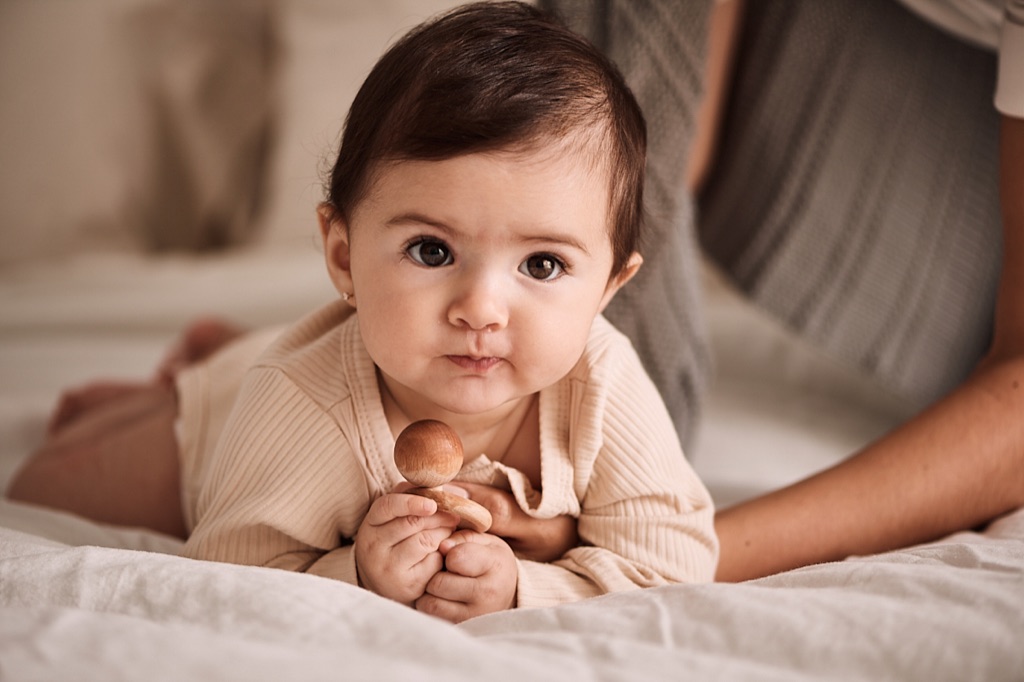 Brown eyed baby on its tummy on a bed clutching a wooden rattle with a parent behind