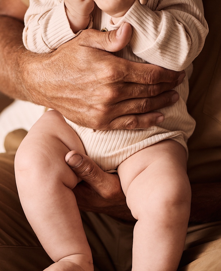 Weathered grandfather hands holding a baby in a cream ribbed onesie on his lap