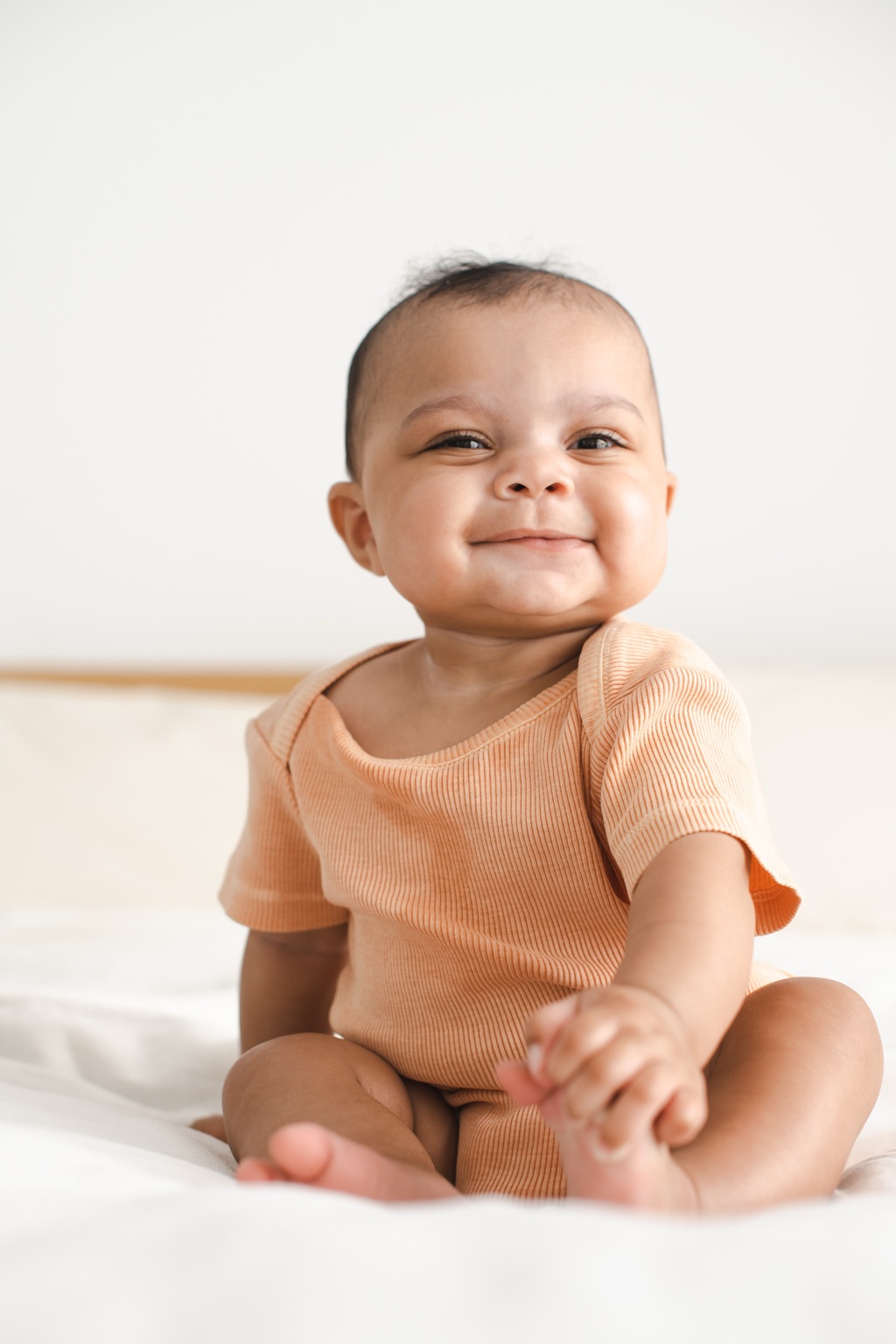 Grinning baby in a ribbed peach onesie sitting on a bed holding its own foot