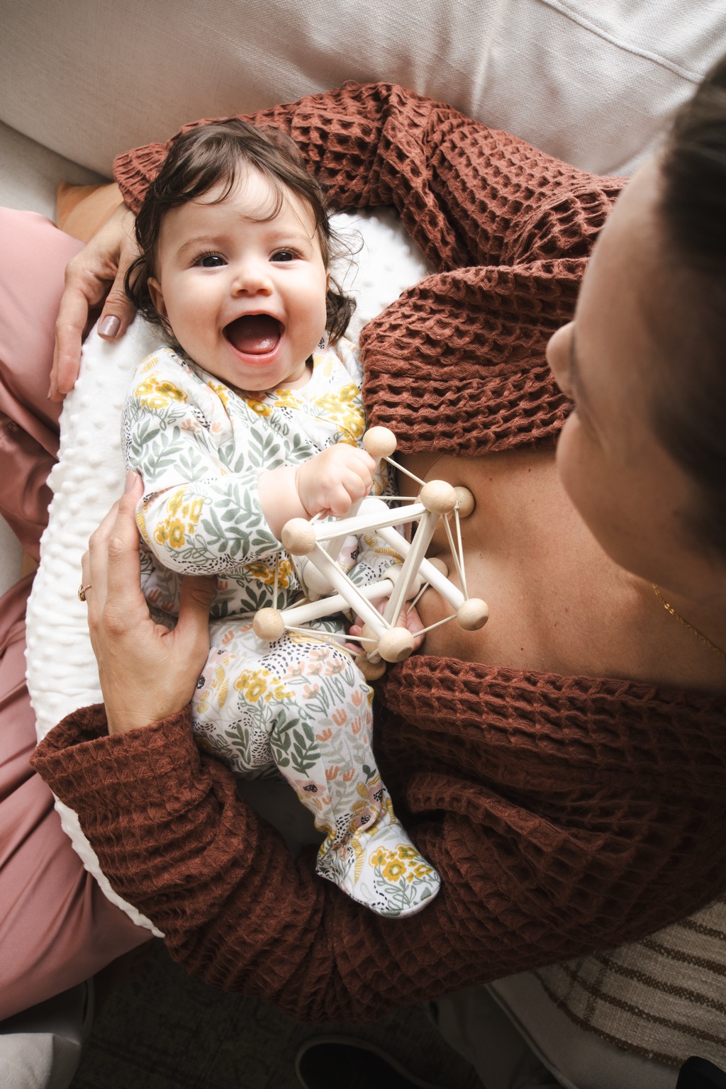 Overhead view of a mom in a rust waffle robe playing with her laughing baby holding a wooden bead rattle