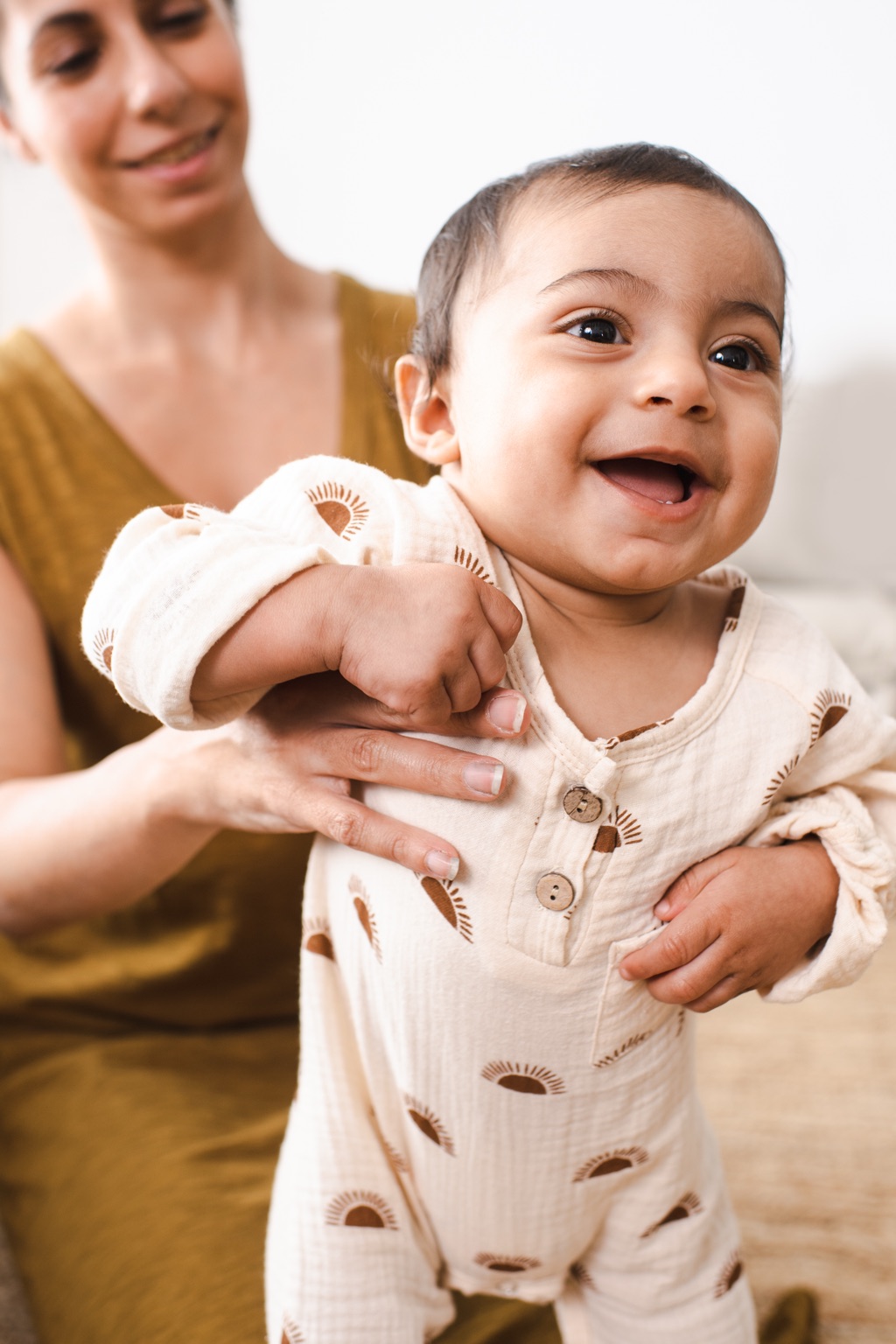 Mom in an olive tank top helping her laughing baby in a sun print muslin romper stand