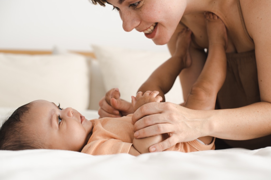 Mom leaning over her baby on the bed, smiling and holding its hands