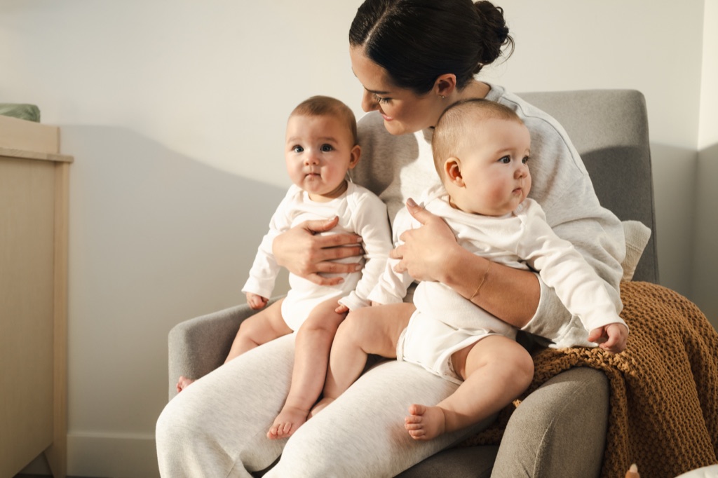Mom holding twin babies in white onesies on a gray armchair