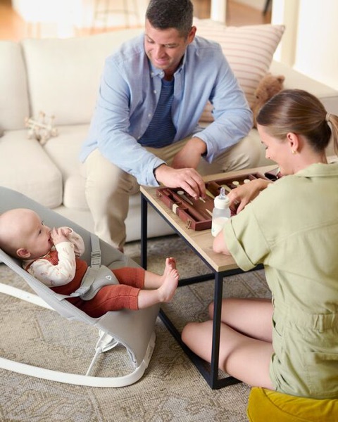 Parents playing backgammon at the coffee table while their baby watches from a bouncer with its bottle