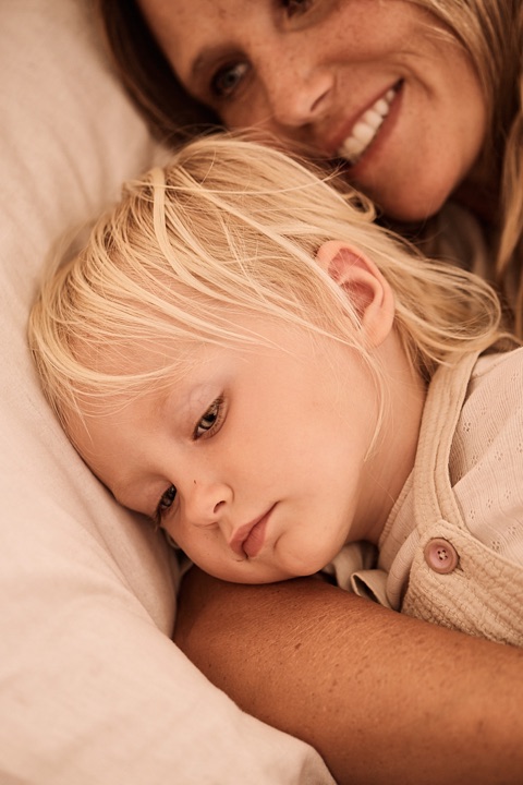 Sleepy blond toddler resting on a pillow with a smiling mom behind