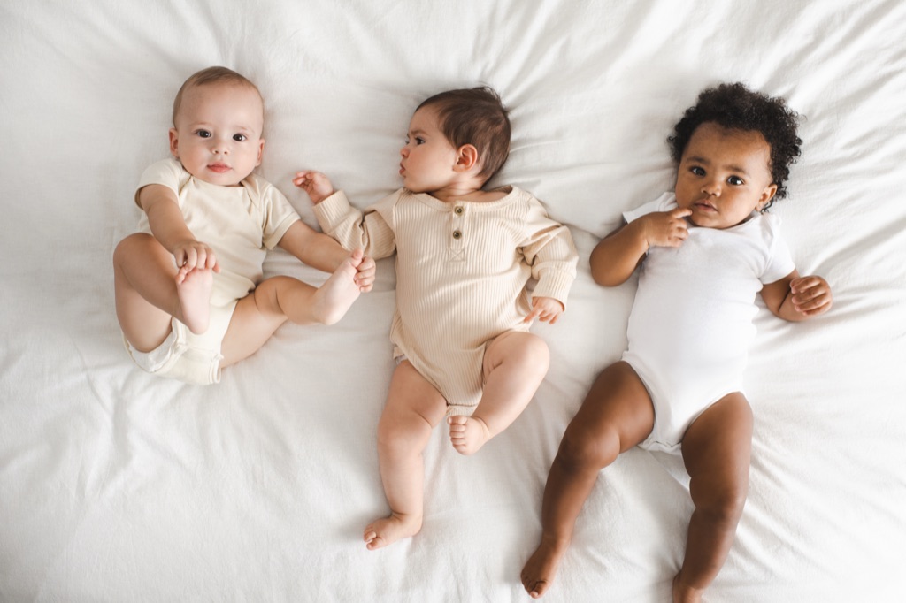 Overhead view of three babies in neutral onesies lying side by side on a white bed