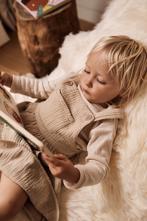 Blond toddler in muslin overalls reading a picture book on sheepskin
