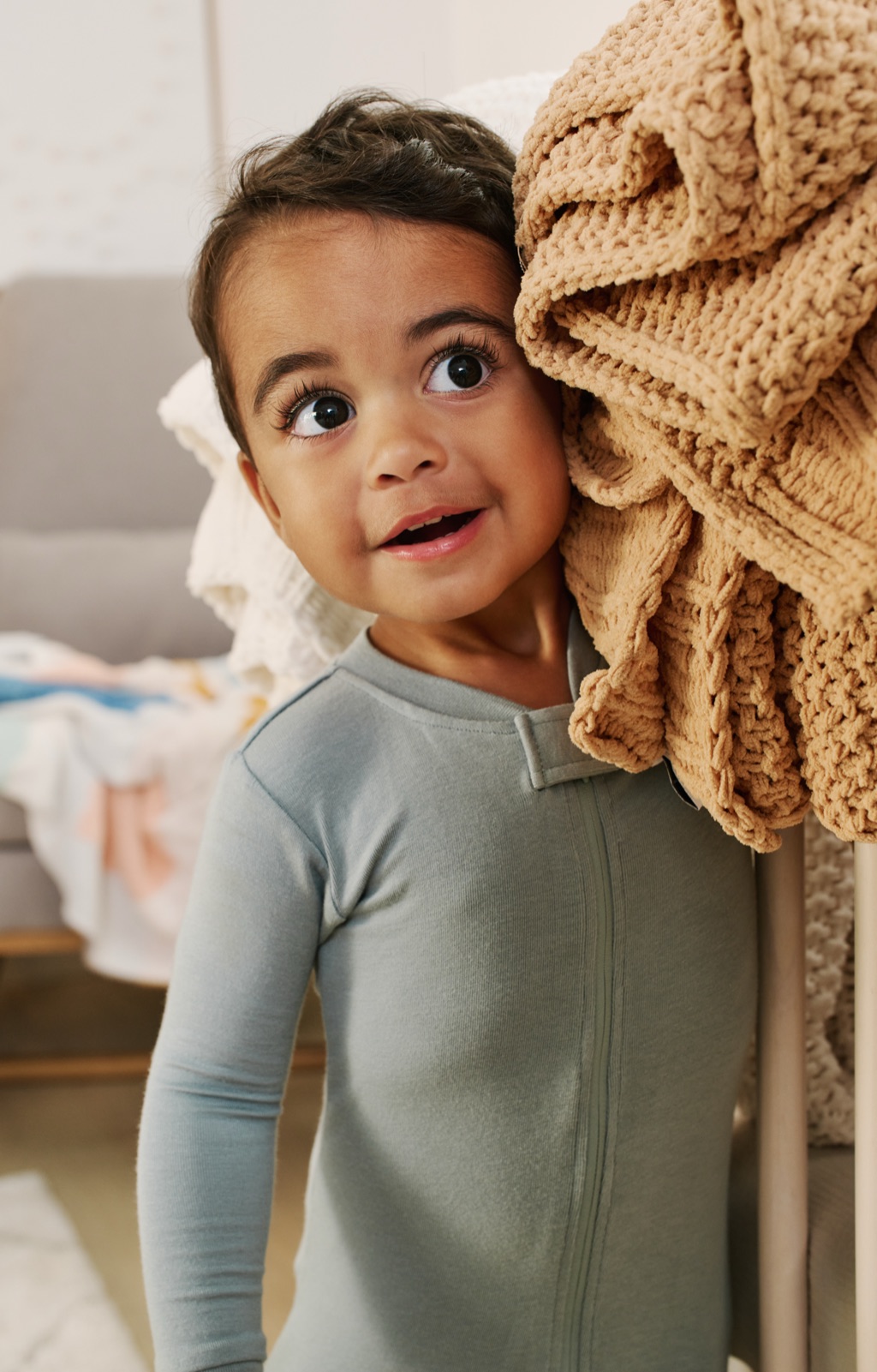 Toddler in sage pajamas peeking around a crib draped with a tan knit blanket