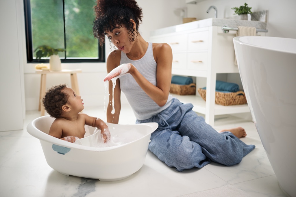 Mom kneeling beside her toddler in a Skip Hop baby tub in a bright bathroom