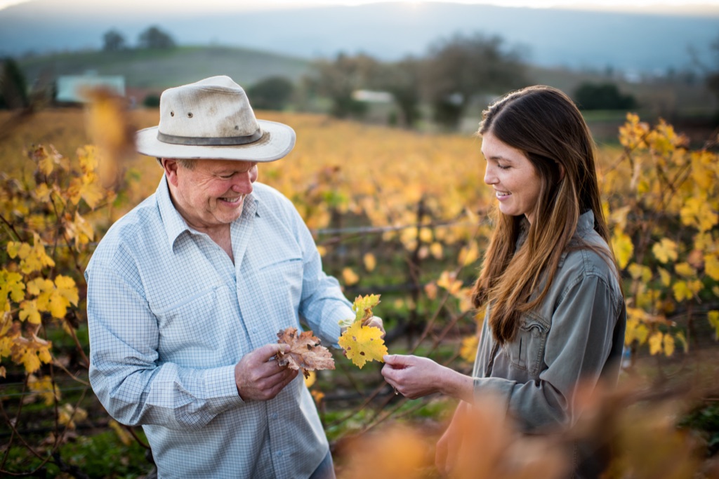 Winegrower and daughter comparing autumn leaves in a golden vineyard at dusk