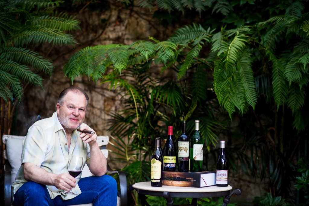 Vintner with a cigar and glass of red beside a lineup of Caymus, Emmolo and Mer Soleil bottles in a fern courtyard