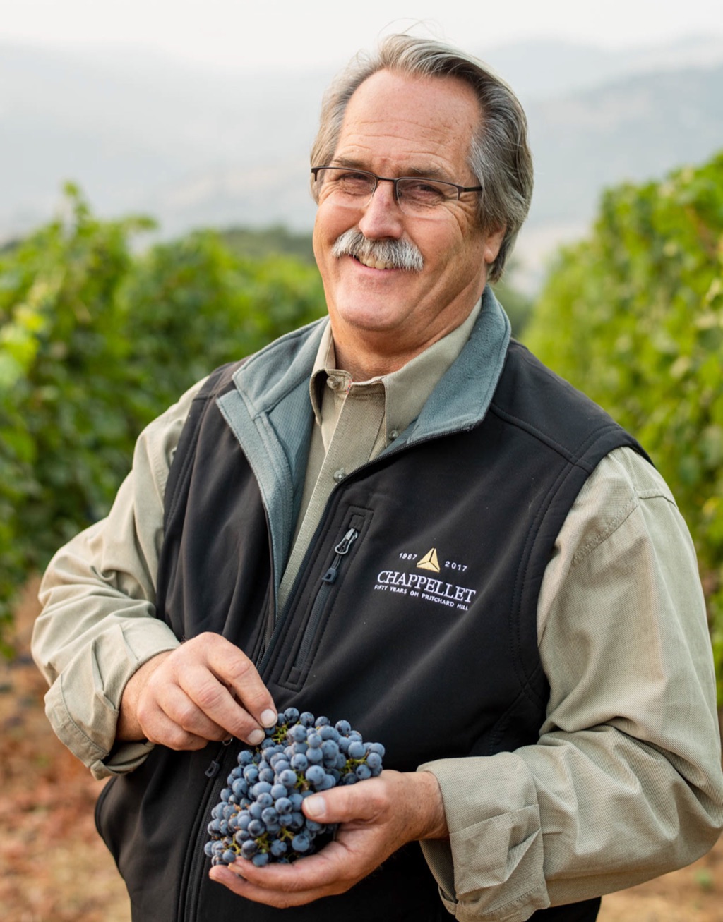 Chappellet vineyard manager smiling with a cluster of cabernet grapes on Pritchard Hill