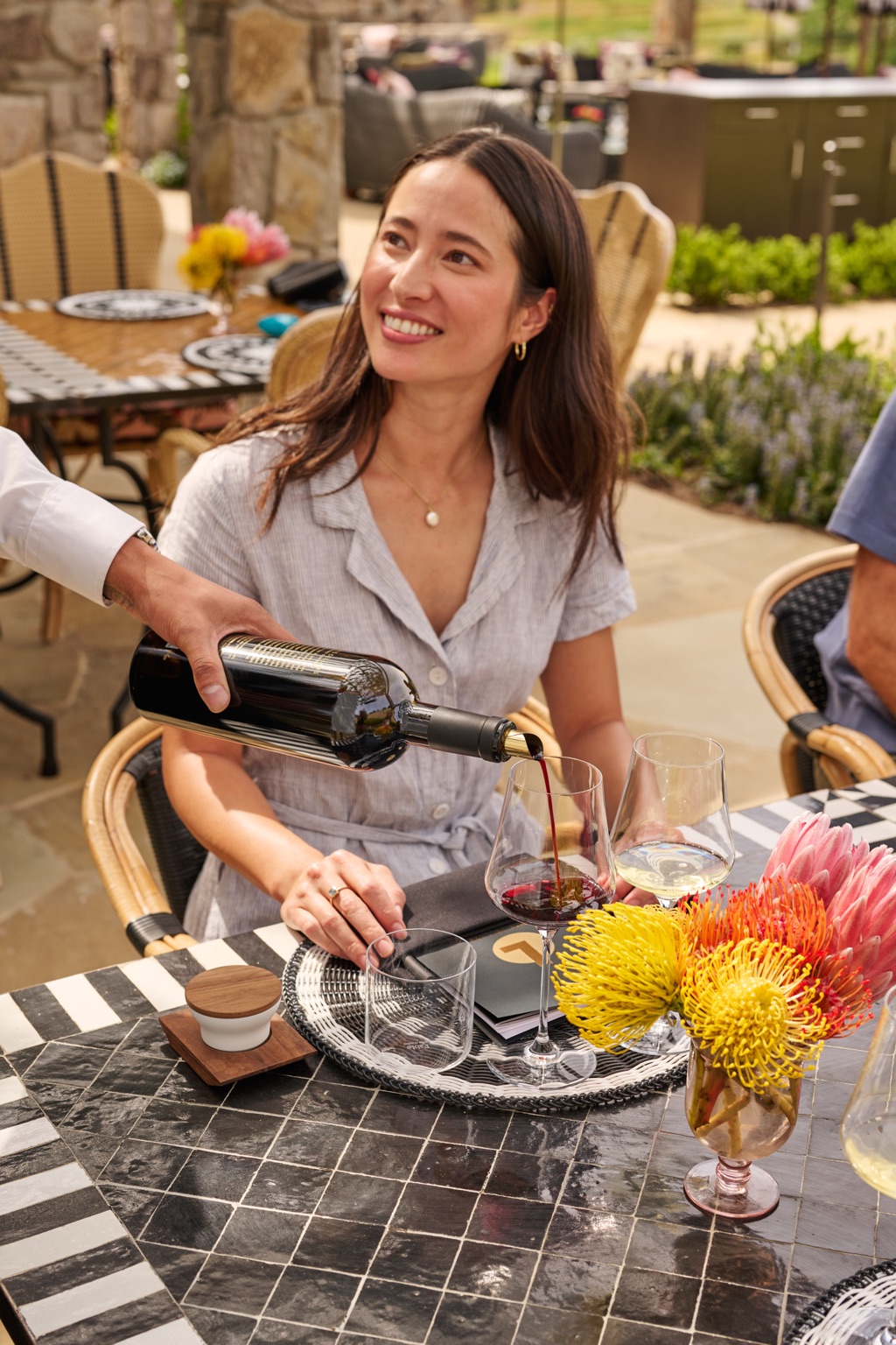 Server pouring red wine tableside for a guest on a Napa Valley winery patio with protea flowers