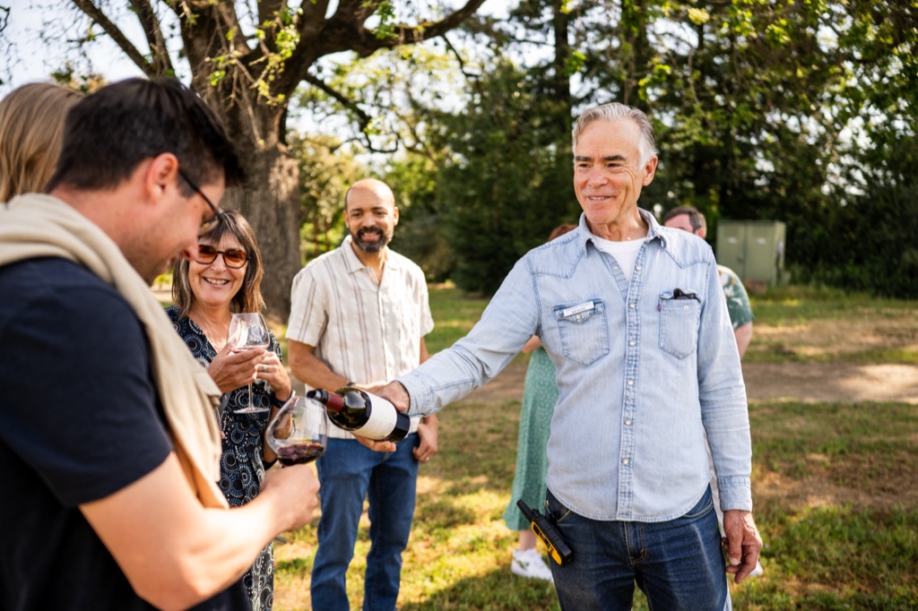 Winery host pouring red wine for a laughing tour group under the oaks