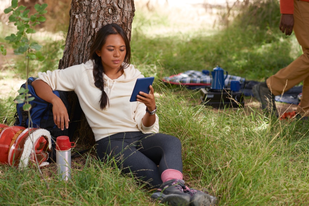 Woman reading a Kindle while leaning against a tree at a campsite, Amazon Kindle lifestyle photography