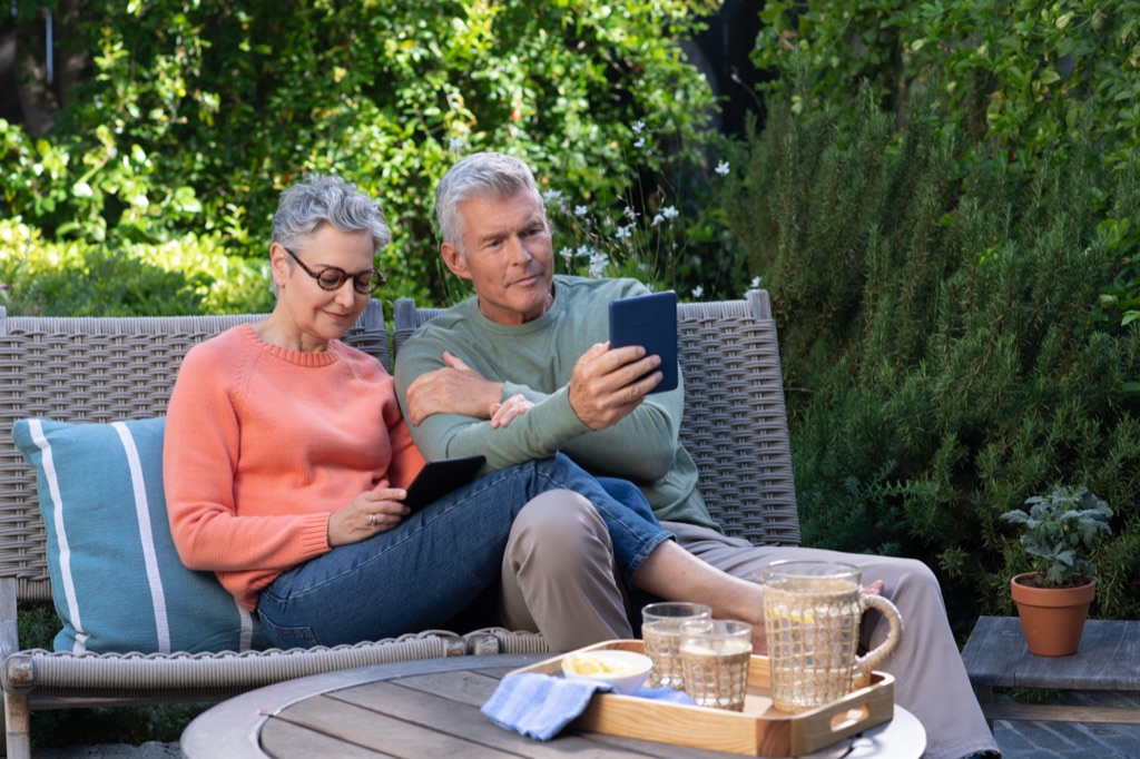 Older couple reading Kindles together on a wicker bench in the garden, Amazon lifestyle photography