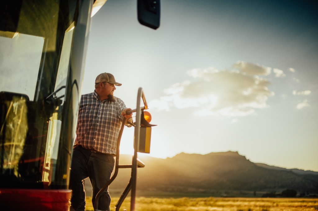 Farmer in plaid and a cap climbing down from a tractor cab at sunset with mountains behind