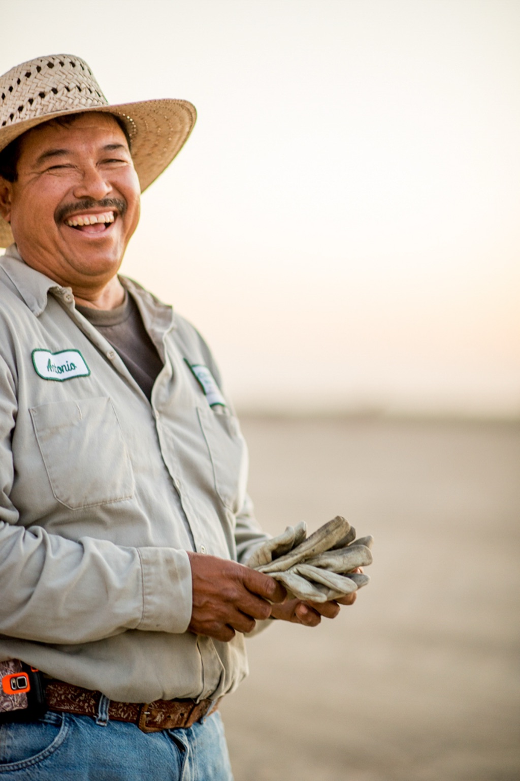 Farmworker in a straw hat laughing at sunset, holding leather work gloves in a hazy field for American AgCredit
