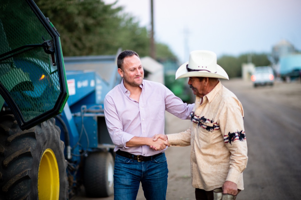 Rancher in a white cowboy hat shaking hands with a younger farmer beside a green tractor