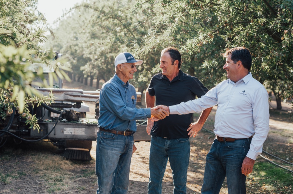 American AgCredit lender shaking hands with two almond growers beside harvest equipment in the orchard