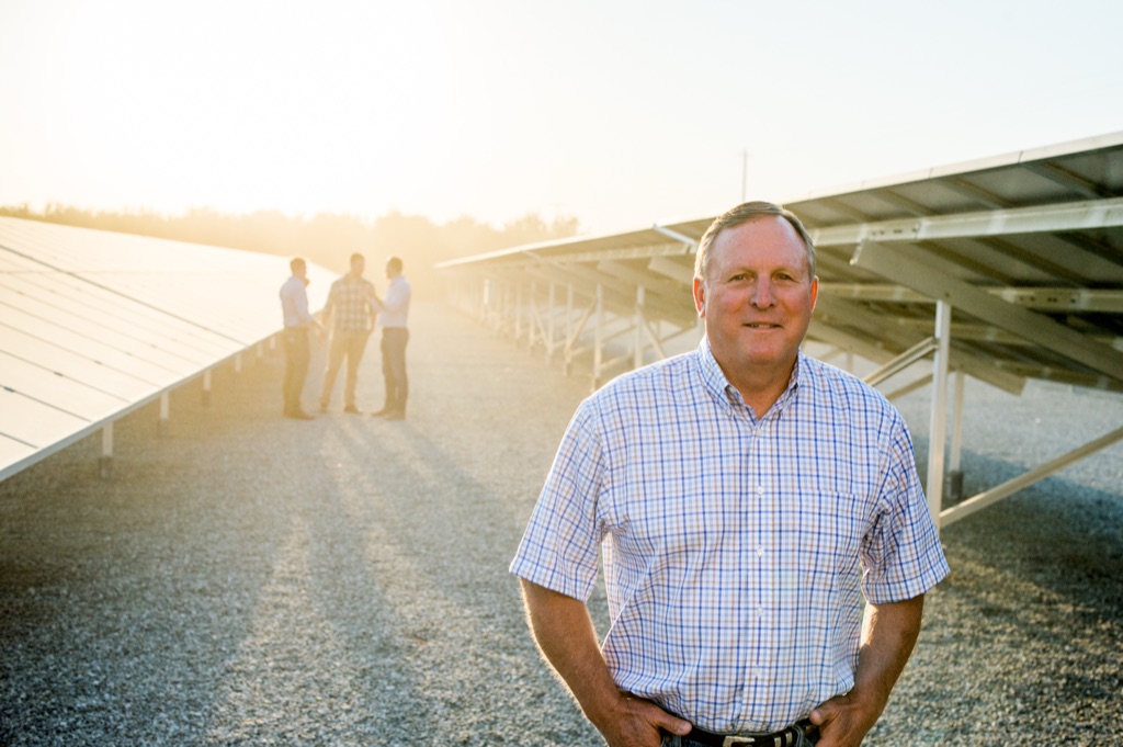 Farmer in a checked shirt at a solar array at sunset, three men talking in the sun flare behind him