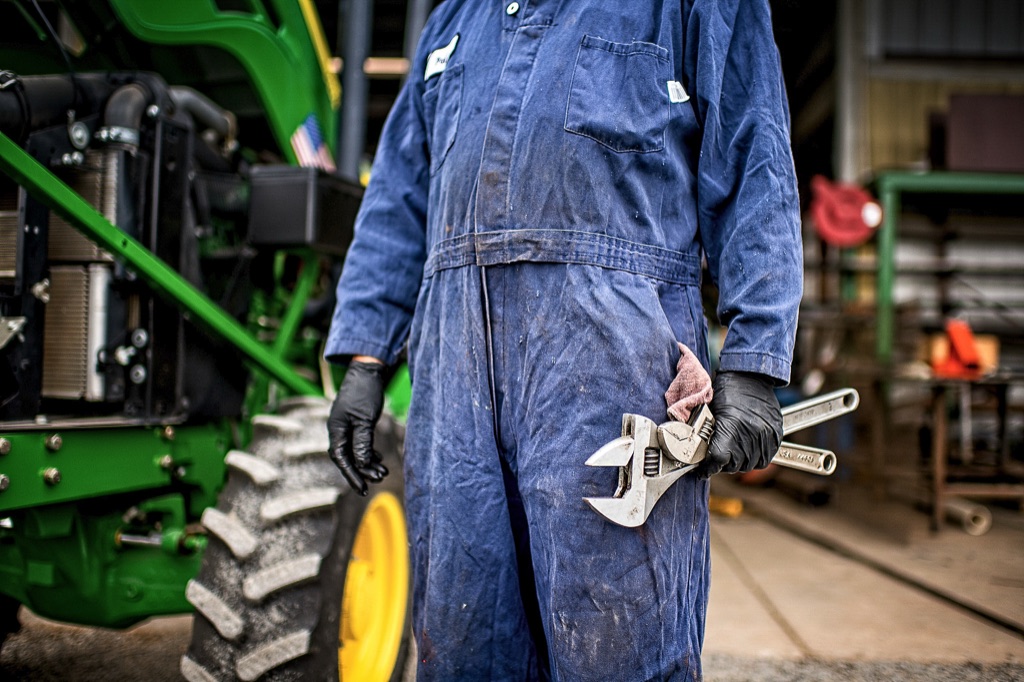Mechanic in blue coveralls holding adjustable wrenches beside a green tractor in the shop