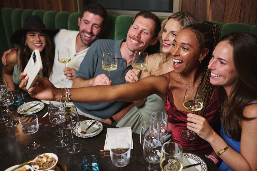 Six friends taking a phone selfie with glasses of white wine in the green velvet banquette