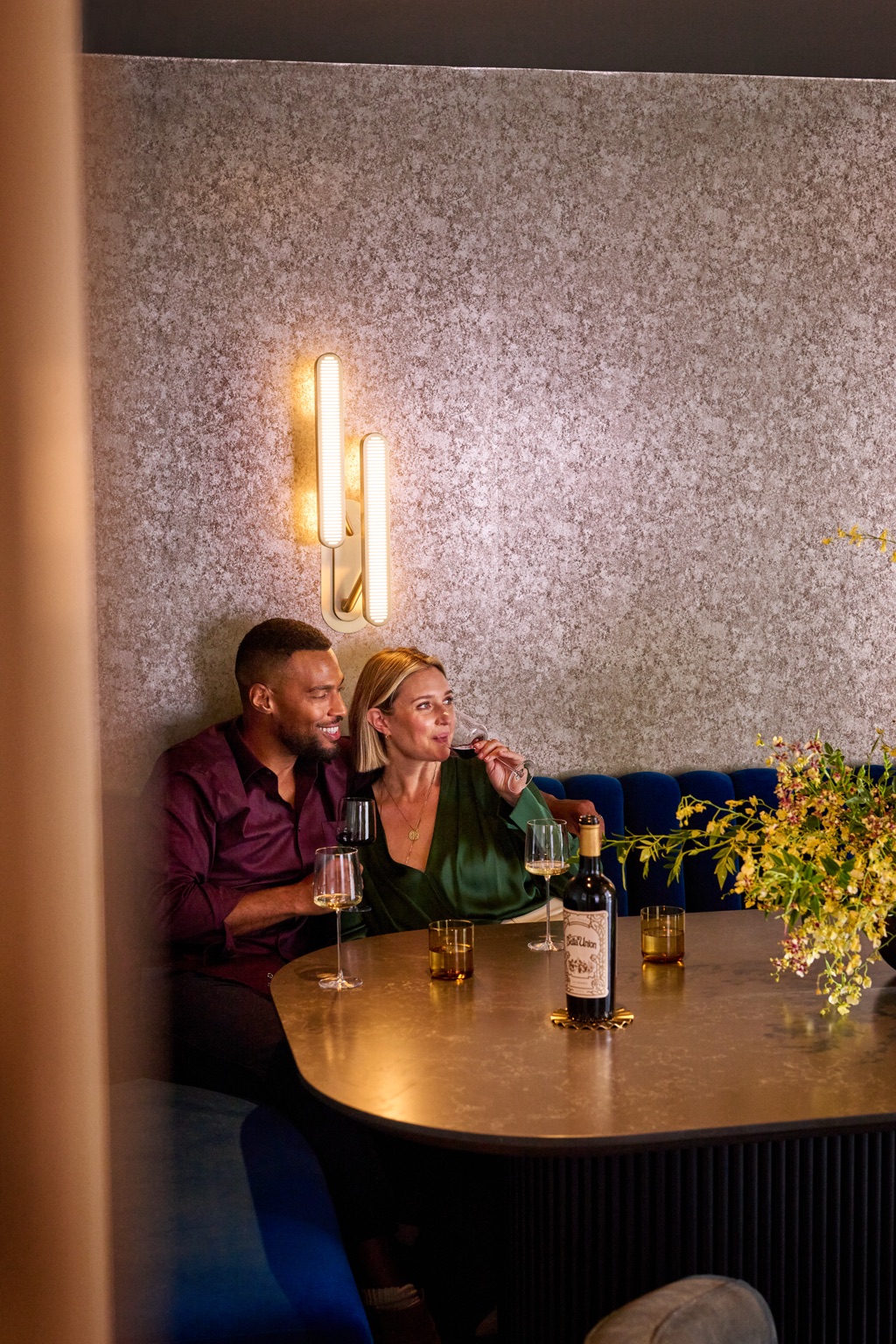 Couple cuddled up in a blue velvet banquette under a brass sconce with a bottle of Bella Union wine
