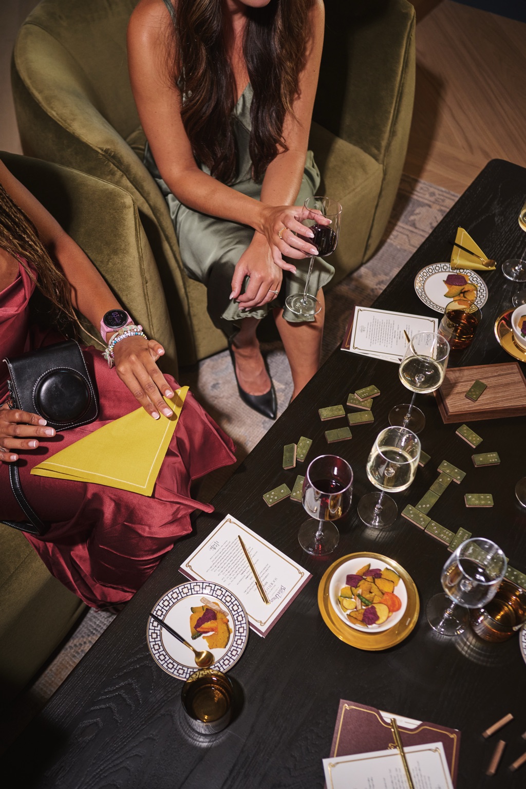 Overhead view of two women playing green leather dominoes with wine, menus, and an instant camera