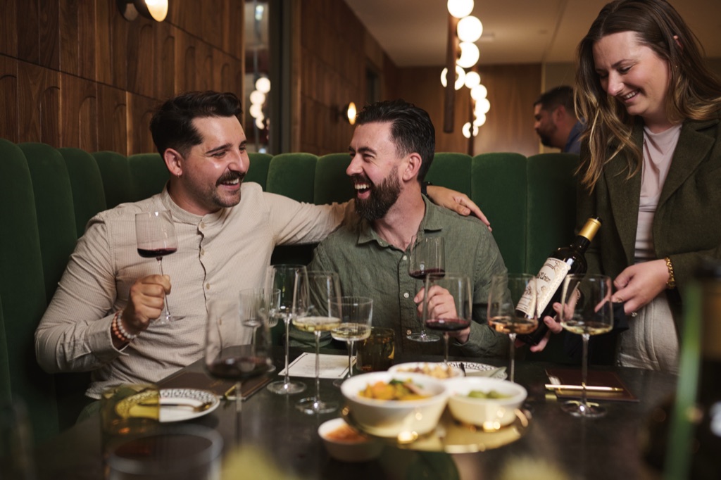 Two men laughing arm in arm in the green banquette as a woman presents a bottle of Bella Union wine