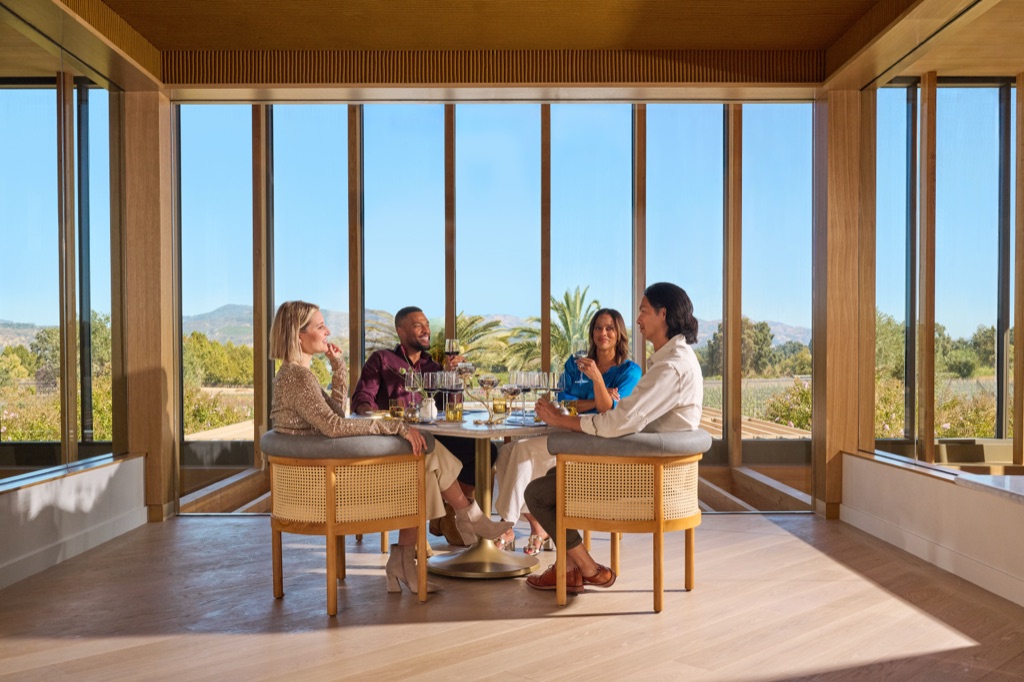 Four friends tasting wine in a glass-walled corner room with cane-back chairs and vineyard views