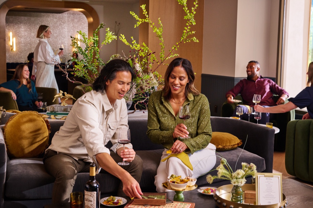 Couple playing mahjong with glasses of red wine on a lounge couch as guests mingle behind them