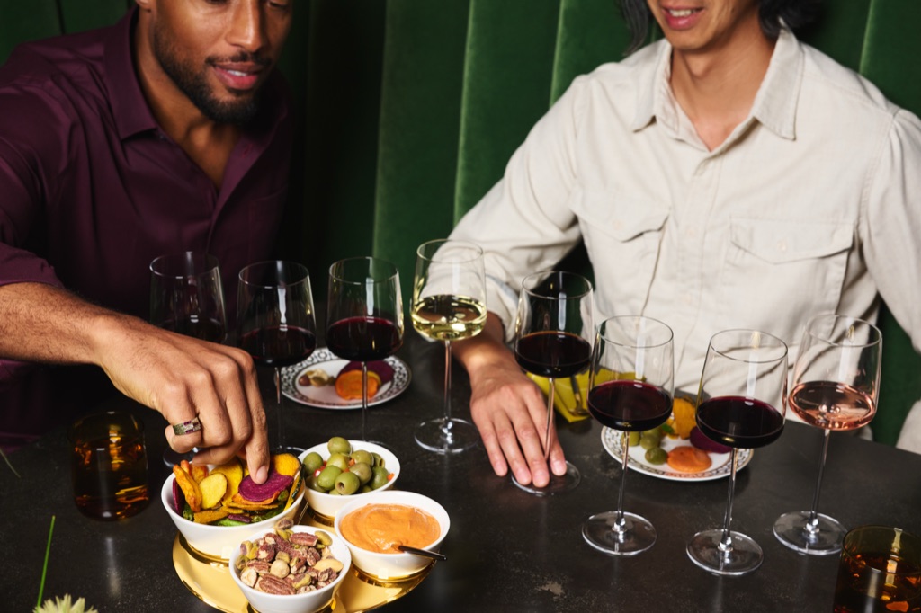 Two men enjoying a seated tasting flight of red, white, and rose wines with a gold snack tray