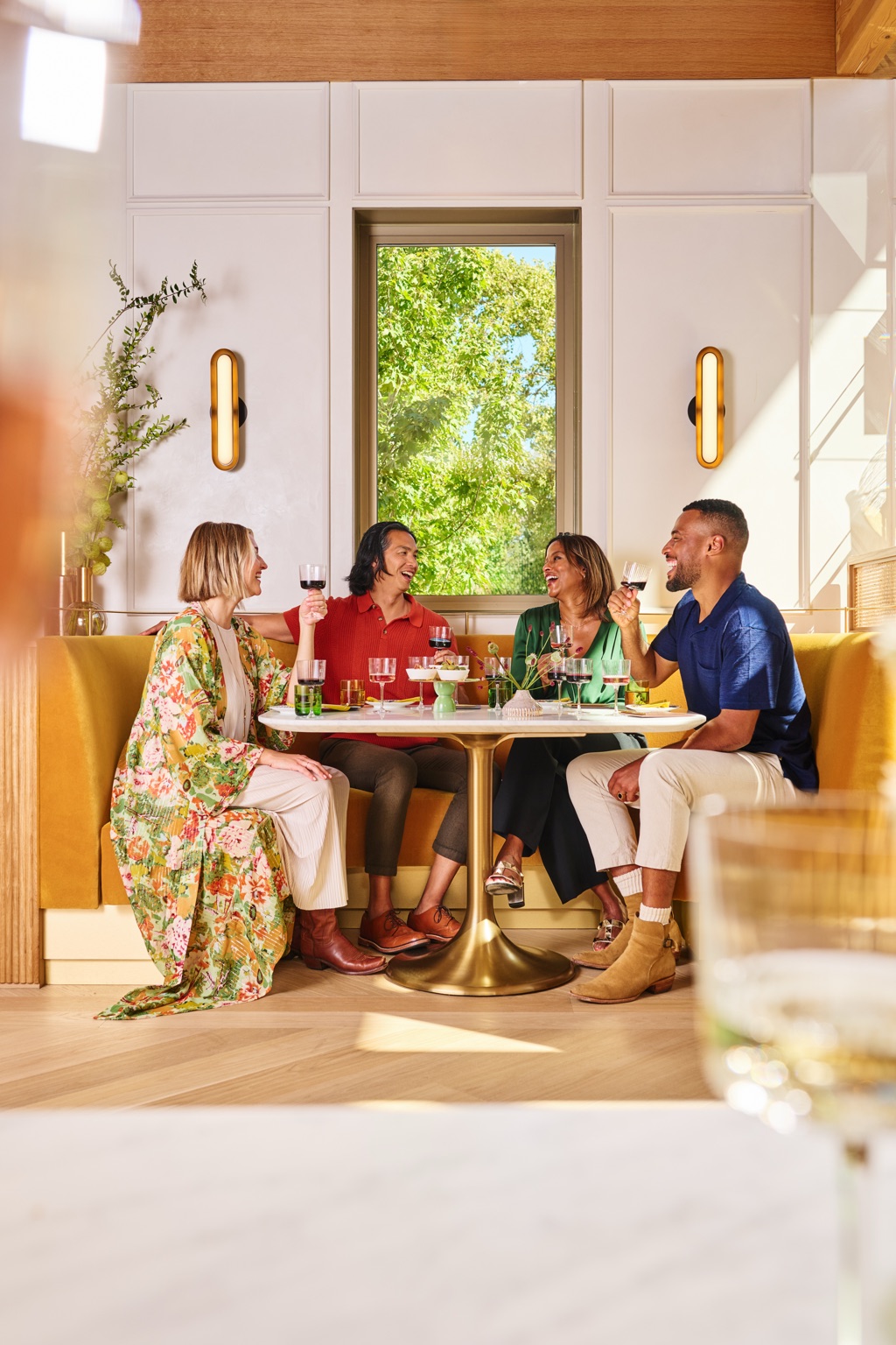 Friends raising red wine over a marble tulip table in a sunlit mustard banquette