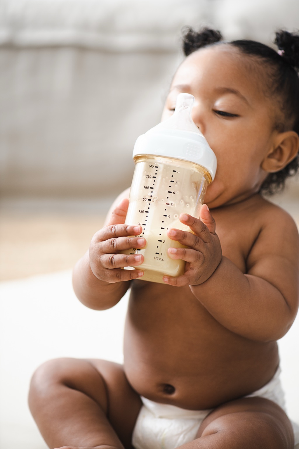 Baby girl holding a measured bottle of ByHeart formula with both hands while drinking