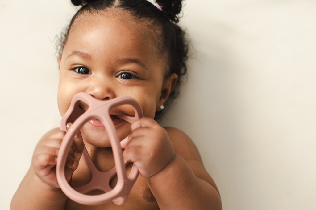 Smiling baby girl with curly pigtails chewing on a pink silicone teether, ByHeart photography