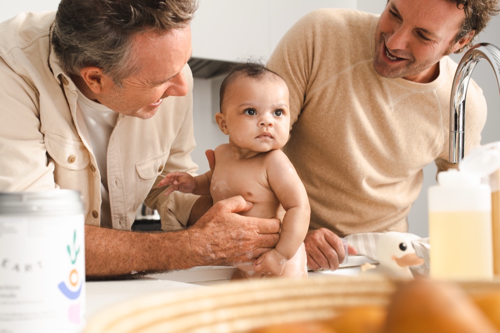 Two dads bathing their baby in the kitchen sink beside a ByHeart formula canister