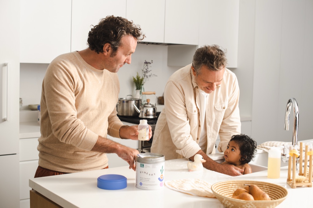 Dad measuring ByHeart formula into a bottle while his partner bathes the baby in the sink