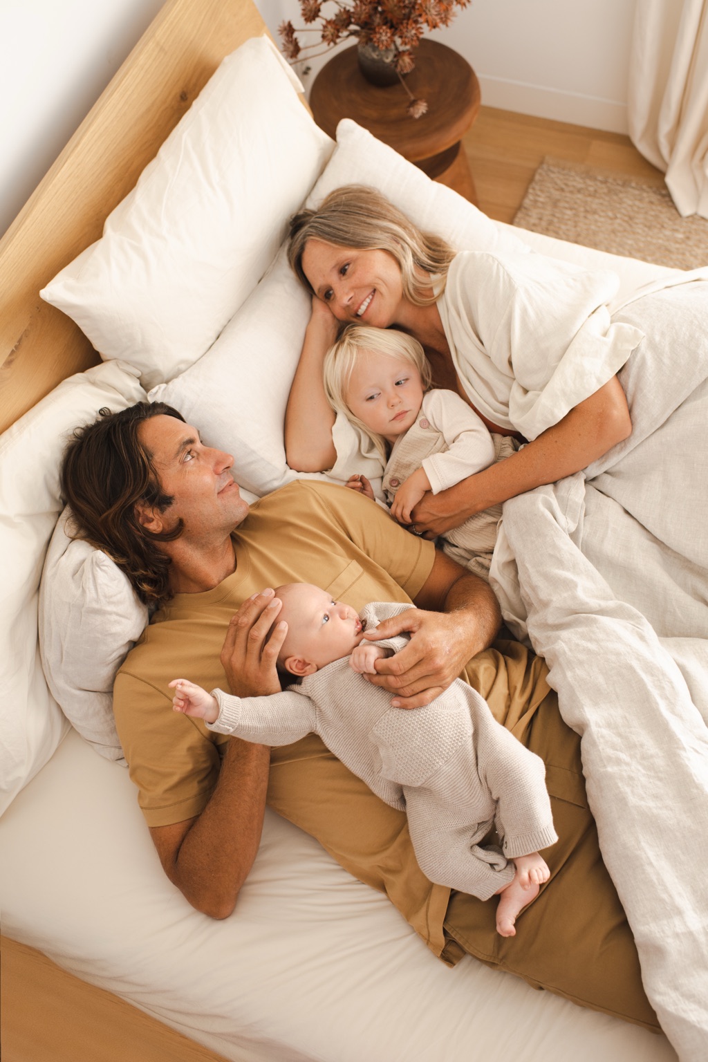 Parents lying in bed with their toddler and newborn in soft morning light, ByHeart campaign photography