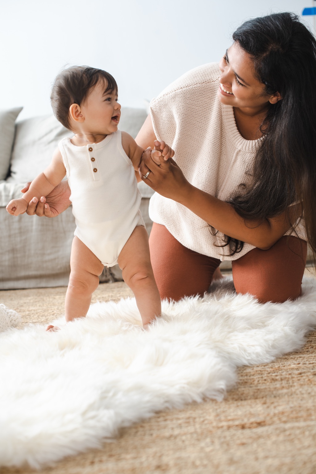 Mother holding her laughing baby's hands as they practice standing on a sheepskin rug