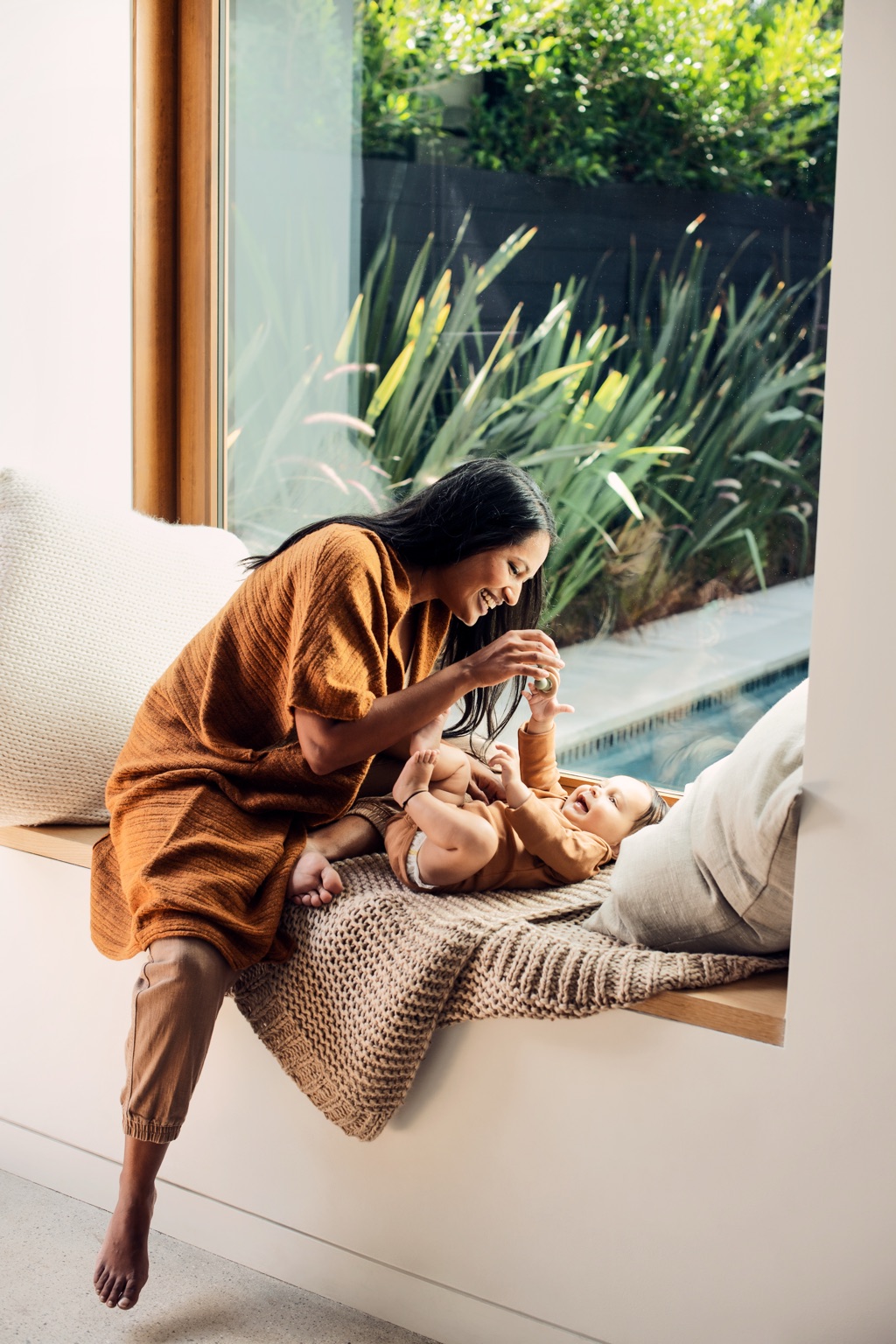 Mother laughing with her baby on a sunny window seat with garden greenery behind, ByHeart lifestyle