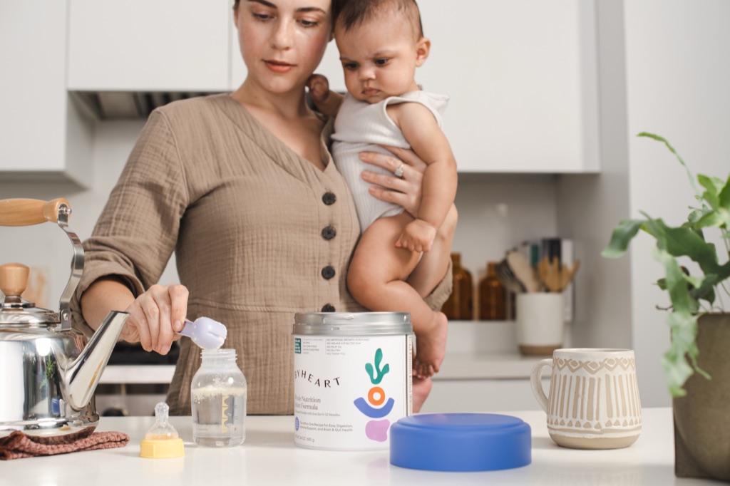 Mother holding her baby on one hip while scooping ByHeart Whole Nutrition Infant Formula into a bottle