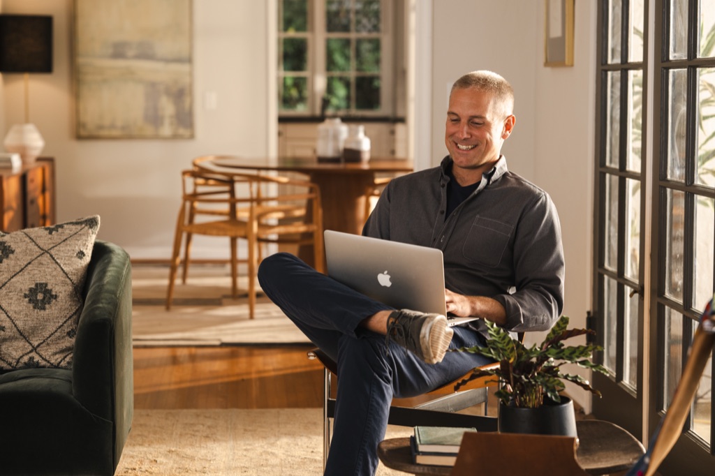 Smiling man working on a laptop in a chair by the french doors in golden afternoon light, photographed by Dawn Heumann
