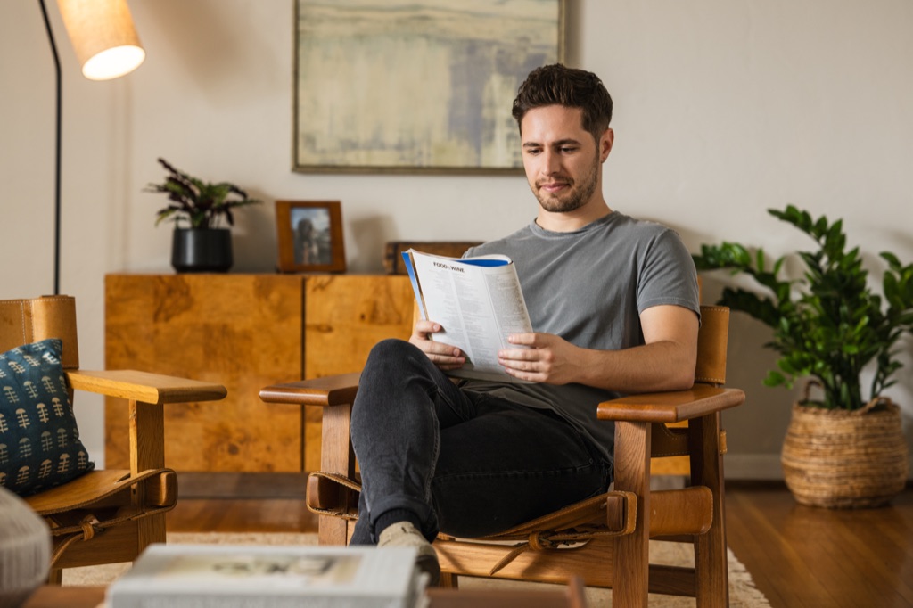 Man reading a magazine in a wood and leather armchair beside a burl cabinet and houseplants