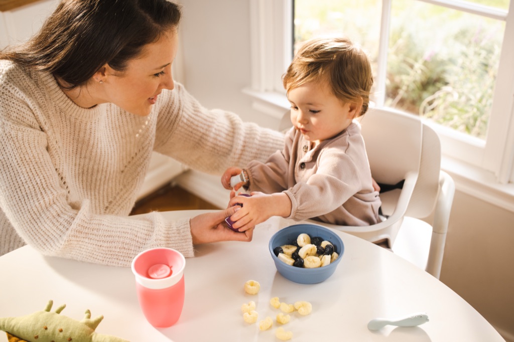 Mom leaning in as her toddler snacks on bananas and blueberries in a high chair by the window