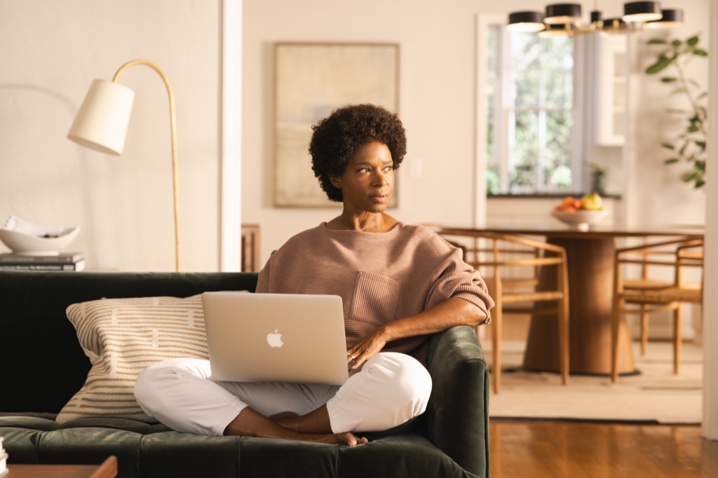 Woman sitting cross-legged on a green velvet sofa with a laptop, gazing out the window