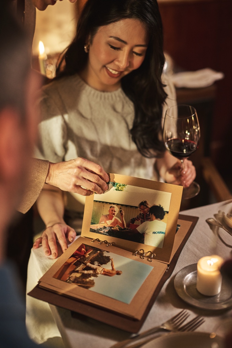 Woman smiling over a photo album of vintage snapshots with a glass of red wine at the table, photographed by Dawn Heumann